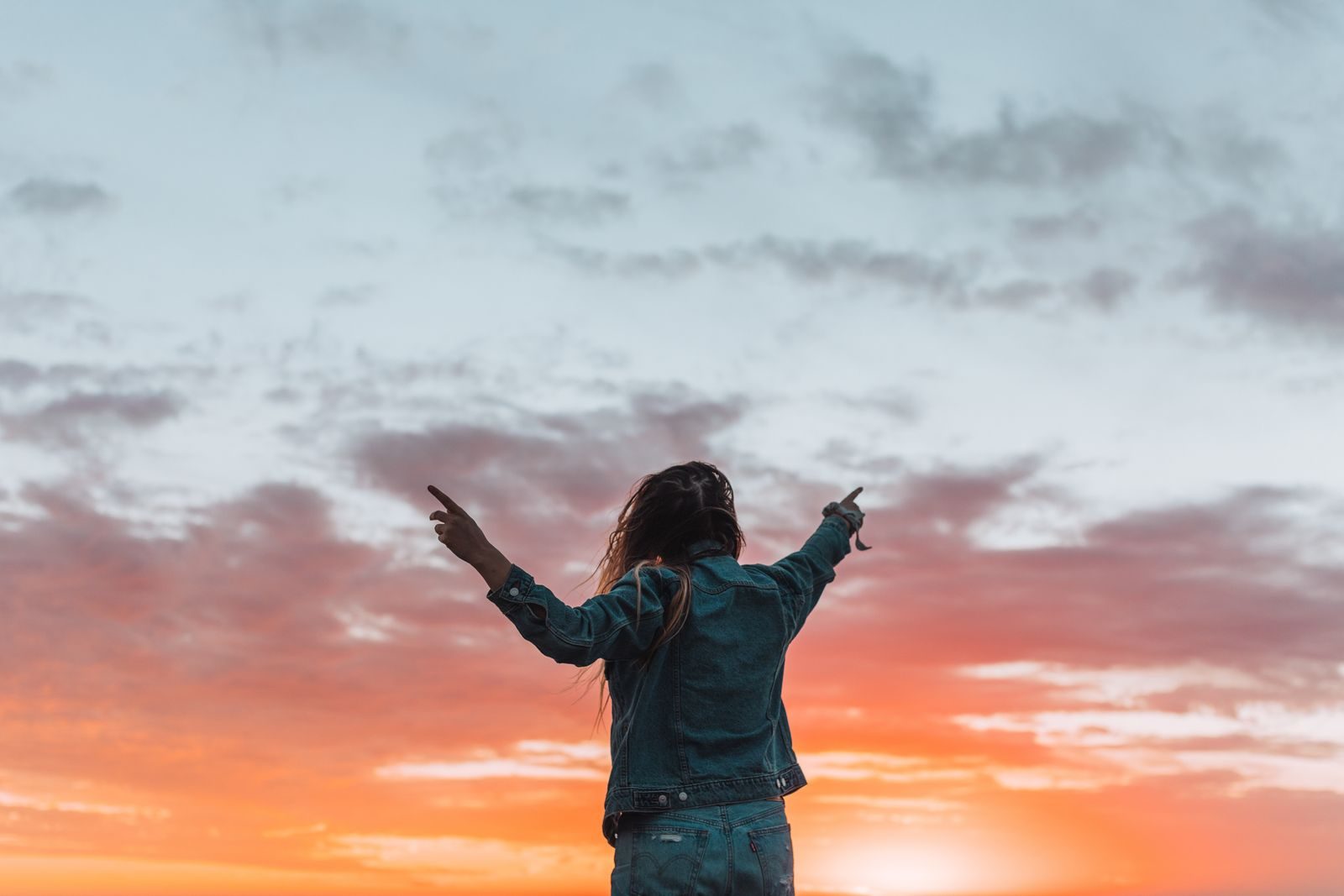 Back view of anonymous female in denim apparel showing colorful cloudy sky with fingers while contemplating sundown