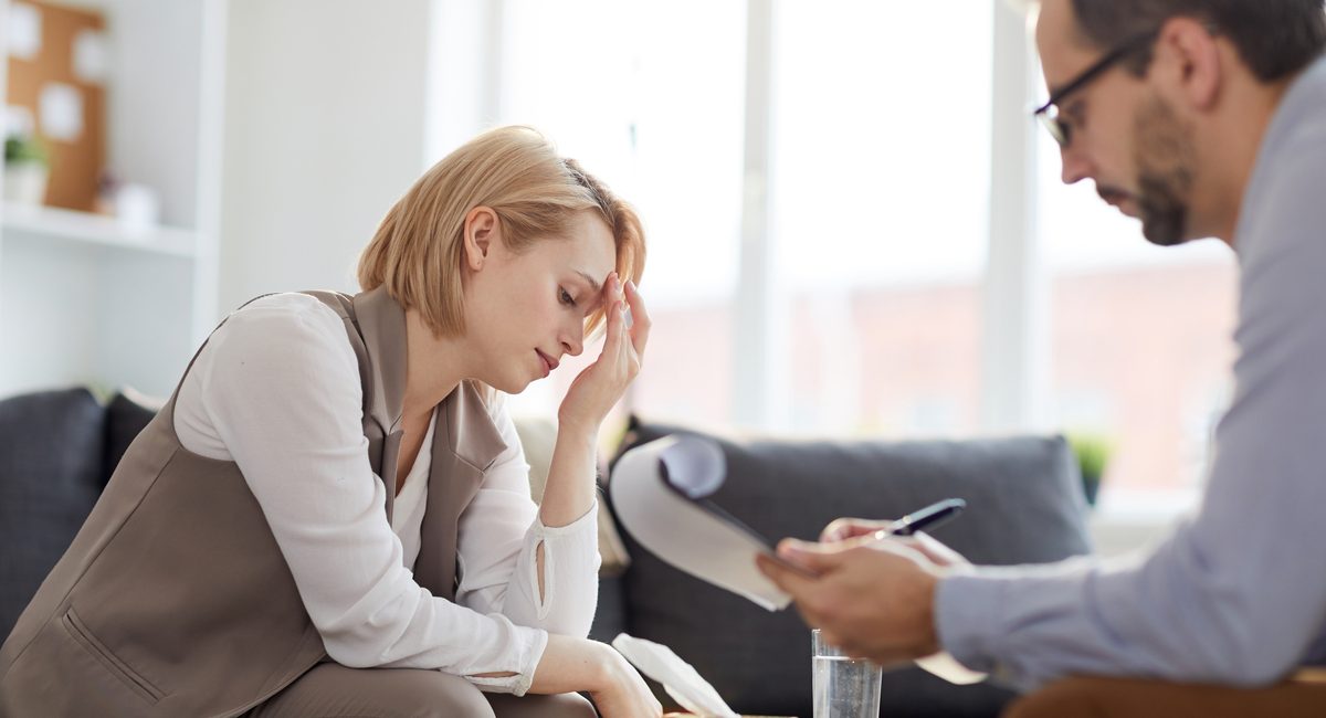 Tired and stressed female touching her forehead while sitting on couch during psychological consultation